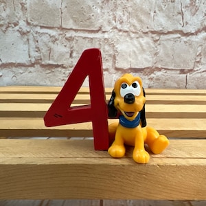 Puede incluir: Un número cuatro de madera rojo con una figurita de perro de dibujos animados amarillo de pie a su lado. El perro tiene ojos negros, una nariz negra y un collar azul.
