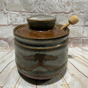 May include: A cylindrical ceramic honey pot with a speckled grey and brown glaze, featuring a matching lid and a light-coloured wooden honey dipper resting inside. The exterior displays a horizontal band of brown decorative markings.