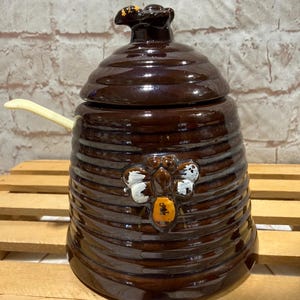 May include: A dark brown glazed ceramic honey pot shaped like a beehive with a matching lid. The pot features a raised bee design with white and orange accents on the front and includes a small, light-colored plastic spoon inserted into the side.