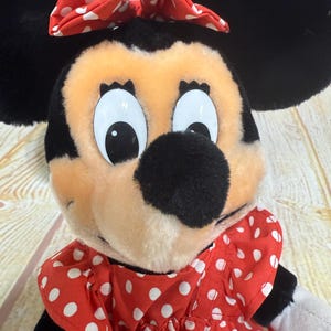 K&ouml;nnte beinhalten: Eine Minnie Mouse Pl&uuml;schpuppe mit schwarzen Ohren, einer schwarzen Nase und einer roten Schleife mit wei&szlig;en Tupfen. Sie tr&auml;gt ein rotes Kleid mit wei&szlig;en Tupfen. Die Puppe hat gro&szlig;e, wei&szlig;&auml;ugige Augen und ein pfirsichfarbenes Gesicht.