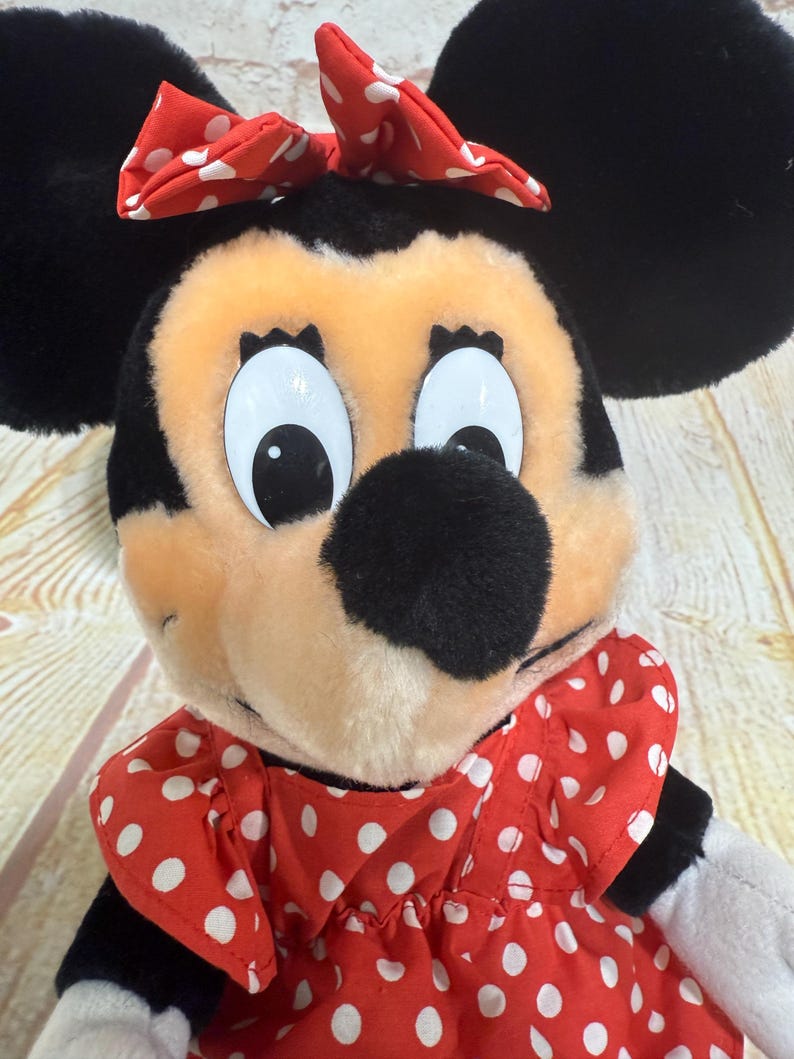 K&ouml;nnte beinhalten: Eine Minnie Mouse Pl&uuml;schpuppe mit schwarzen Ohren, einer schwarzen Nase und einer roten Schleife mit wei&szlig;en Tupfen. Sie tr&auml;gt ein rotes Kleid mit wei&szlig;en Tupfen. Die Puppe hat gro&szlig;e, wei&szlig;&auml;ugige Augen und ein pfirsichfarbenes Gesicht.