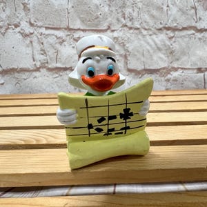 May include: A white duck figurine wearing a green shirt and a white hat, holding a yellow scroll with black musical notes.