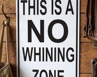 Op de afbeelding: Een rechthoekig wit bord met zwarte tekst: "THIS IS A NO WHINING ZONE." Het bord is bevestigd aan een houten muur met een zwarte rand en wordt op zijn plaats gehouden door twee bruine schroeven.