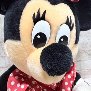 May include: Close-up of a Minnie Mouse plush toy. The toy has a black head and ears, a beige face, and large white eyes with black pupils. A red bow with white polka dots sits atop the head, and a matching bow is around the neck.