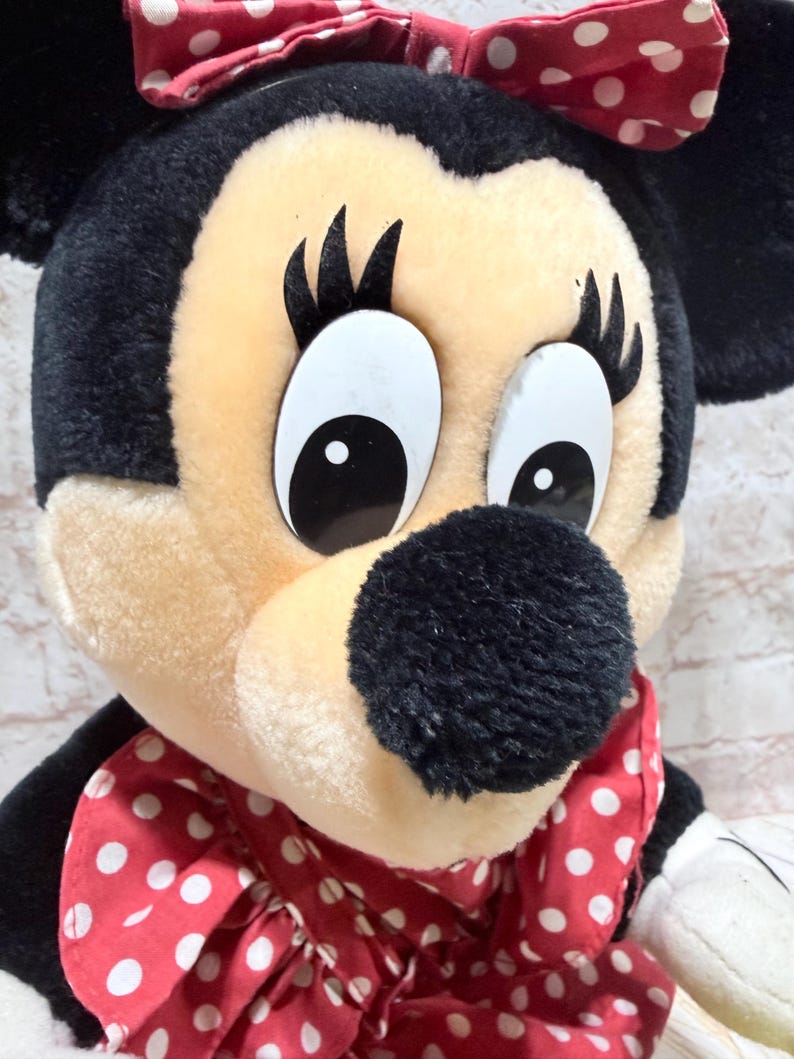 May include: Close-up of a Minnie Mouse plush toy. The toy has a black head and ears, a beige face, and large white eyes with black pupils. A red bow with white polka dots sits atop the head, and a matching bow is around the neck.
