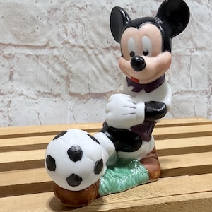 Pu&ograve; includere: Una statuina di Topolino in uniforme da calcio, in posa con un pallone da calcio. La statuina &egrave; principalmente bianca, nera e marrone, con un colletto viola. Il pallone da calcio &egrave; bianco con pentagoni neri, su una base verde.