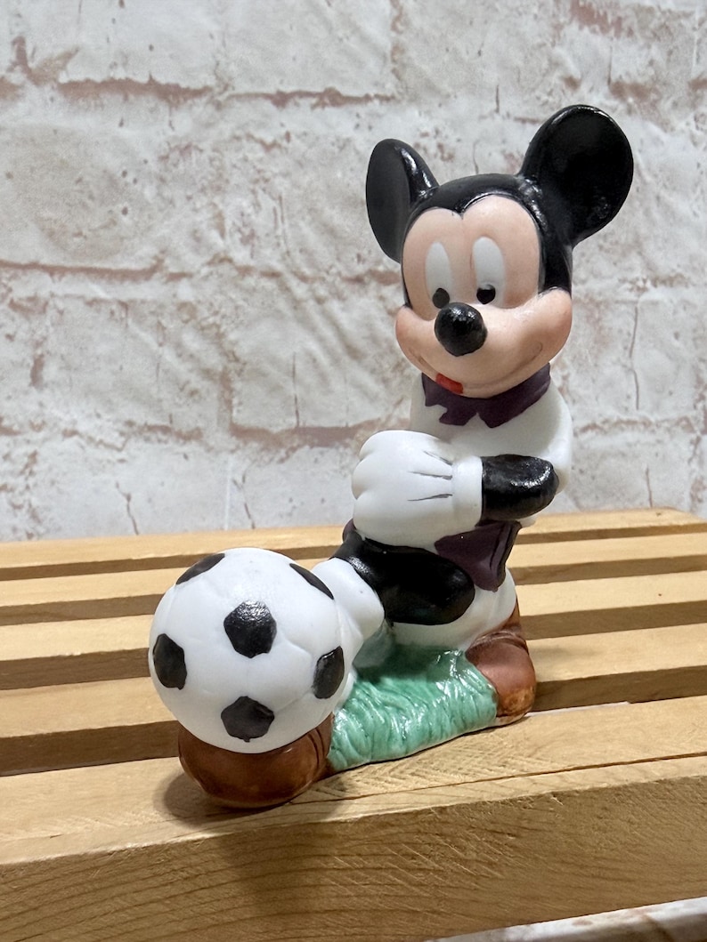 Pu&ograve; includere: Una statuina di Topolino in uniforme da calcio, in posa con un pallone da calcio. La statuina &egrave; principalmente bianca, nera e marrone, con un colletto viola. Il pallone da calcio &egrave; bianco con pentagoni neri, su una base verde.