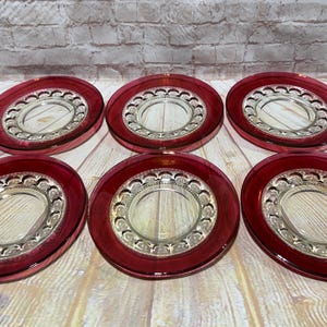Op de afbeelding: Een set van zes ronde glazen borden met een dieprode buitenrand en een helder middengedeelte. Elk bord heeft een decoratief reliëfpatroon met een reeks kleine, herhalende cirkelvormige uitsparingen rondom het centrale gedeelte.