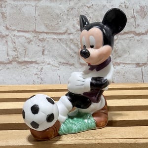 Pu&ograve; includere: Una statuetta in ceramica di Topolino in uniforme da calcio, pronto a calciare un pallone da calcio. La statuetta presenta una combinazione di colori bianco e nero con accenti di rosso, marrone e verde. La statuetta &egrave; su una superficie di legno.