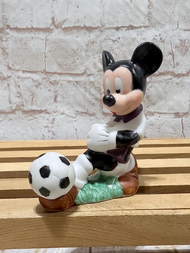 Pu&ograve; includere: Una statuetta in ceramica di Topolino in uniforme da calcio, pronto a calciare un pallone da calcio. La statuetta presenta una combinazione di colori bianco e nero con accenti di rosso, marrone e verde. La statuetta &egrave; su una superficie di legno.