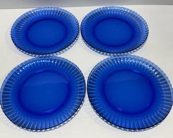 May include: Four cobalt blue glass plates. Each plate has a ribbed rim and a smooth, deep blue center. The plates are arranged in a square formation, showcasing their matching design and color.