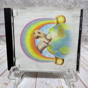 Op de afbeelding: Een cd-hoesje met een kleurrijke illustratie. Het kunstwerk bevat een regenboog, een wereldbol en een cartoonachtig personage. De hoes wordt gepresenteerd op een transparante plastic standaard, tegen een lichte houten ondergrond.