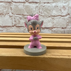 Peut inclure: Une petite figurine rose et grise d'une souris de dessin animé portant un nœud rose. La souris est debout, les bras croisés, et a une expression joyeuse sur son visage.