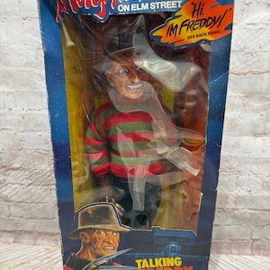 May include: A vintage Matchbox "A Nightmare on Elm Street" Talking Freddy Krueger doll in its original packaging. The box features the film's title, Freddy's image, and the phrase "Hi, I'm Freddy!" The doll is wearing a striped sweater.