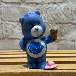May include: A blue Care Bear figurine with a cloud and rain design on its belly. The bear is holding a brown object and wearing a pink hat. The figurine has a white muzzle and a blue nose.