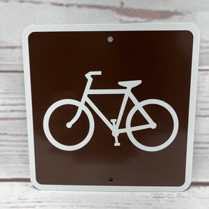 May include: Brown metal sign with a white bicycle symbol. The sign has two holes for hanging.