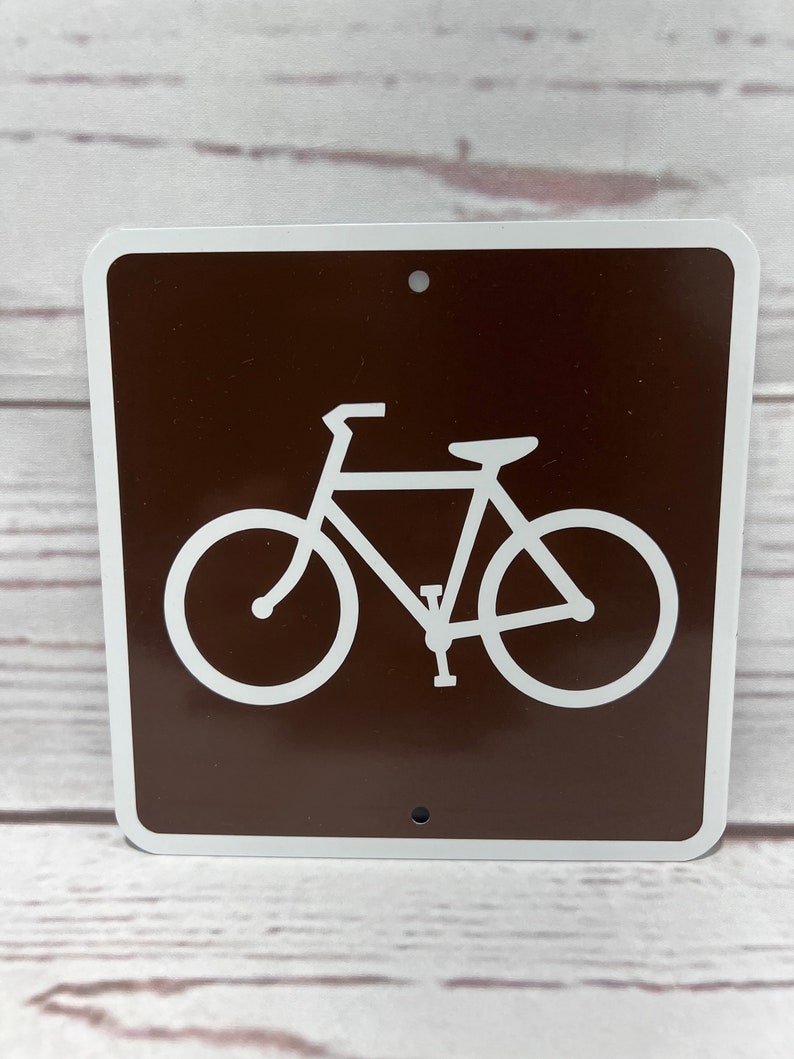 May include: Brown metal sign with a white bicycle symbol. The sign has two holes for hanging.