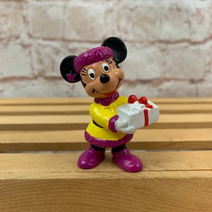 May include: A purple and yellow Christmas-themed figurine of Minnie Mouse holding a white gift box with a red ribbon.