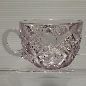 May include: A clear, lavender-tinted glass teacup with a handle. The cup features a diamond-cut pattern across its surface. The handle is smooth and rounded, and the cup sits on a clear, flat surface.