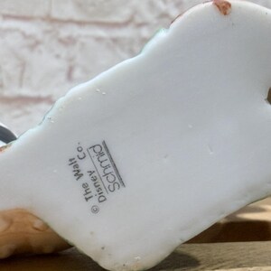 Pu&ograve; includere: Una statuetta in ceramica bianca con dettagli verdi e marroni. La base della statuetta riporta la scritta "The Walt Disney Co. Schmid". La statuetta &egrave; su una superficie di legno.