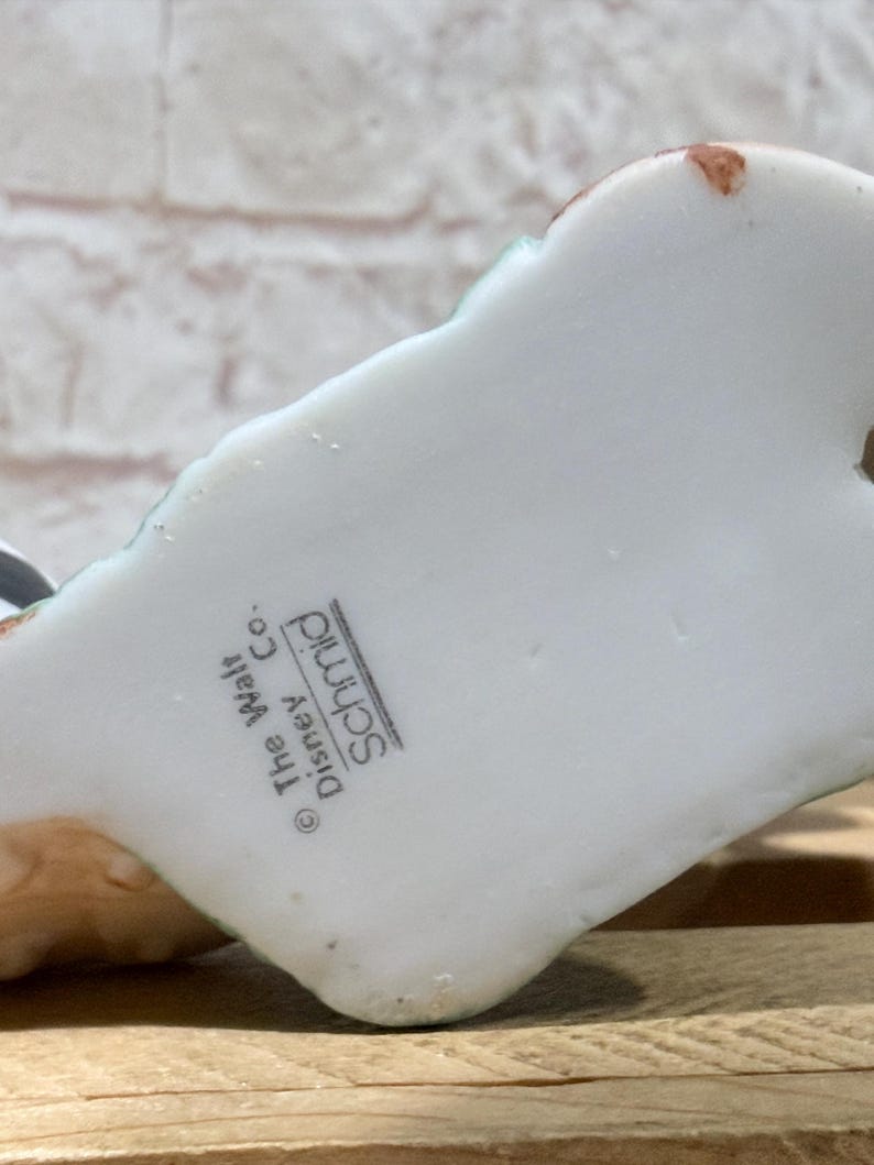 Pu&ograve; includere: Una statuetta in ceramica bianca con dettagli verdi e marroni. La base della statuetta riporta la scritta "The Walt Disney Co. Schmid". La statuetta &egrave; su una superficie di legno.