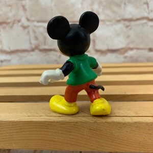 以下が含まれることがあります： 緑のシャツ、赤いズボン、黄色の靴を履いたミッキーマウスの黒と白のプラスチック製フィギュア。フィギュアは木製の表面に立っています。
