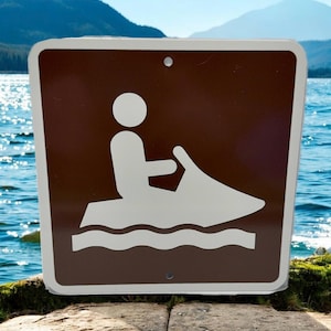 May include: A brown and white sign depicting a person on a jet ski. The sign has a white border and is set against a backdrop of water and mountains. The sign is likely a water safety notice.