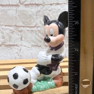 Pu&ograve; includere: Statuetta in ceramica di Topolino in uniforme da calciatore, pronto a calciare un pallone. La statuetta &egrave; principalmente bianca, nera e marrone, con un tocco di verde. La statuetta &egrave; alta circa 7.6 cm.