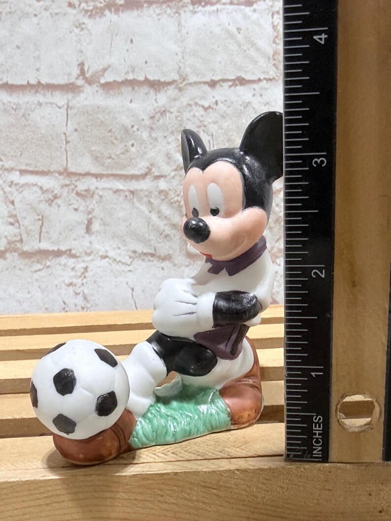 Pu&ograve; includere: Statuetta in ceramica di Topolino in uniforme da calciatore, pronto a calciare un pallone. La statuetta &egrave; principalmente bianca, nera e marrone, con un tocco di verde. La statuetta &egrave; alta circa 7.6 cm.