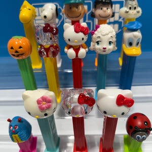 Puede incluir: Una colección de dispensadores PEZ con varios personajes de dibujos animados, incluyendo Hello Kitty y Snoopy. Los dispensadores tienen tallos coloridos y cabezas de personajes, con una variedad de colores y diseños.