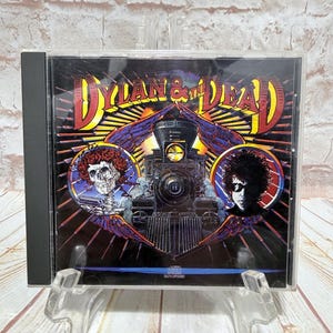 May include: CD case for the album "Dylan & The Dead." The cover art features a train, a skull with roses, and a portrait of a person with sunglasses. The text "Dylan & The Dead" is in yellow and red.