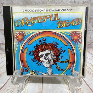 May include: A Grateful Dead CD case featuring a skull with roses, set against a blue background. The case has the text "2 RECORD SET ON 1 SPECIALLY-PRICED DISC" and the band's name in a colorful, psychedelic design. The CD case is displayed on a clear plastic stand.