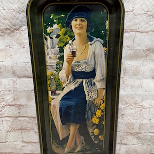 May include: A rectangular metal Coca-Cola serving tray with a black border and gold trim. The center features a vintage illustration of a person in a blue hat and dress holding a glass of soda, with a Coca-Cola bottle nearby and yellow flowers.