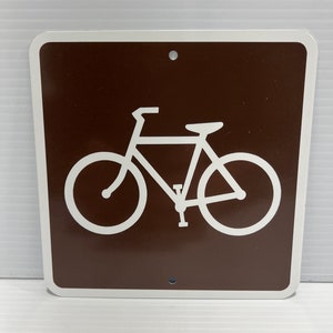 May include: A brown metal sign with a white bicycle symbol. The sign has two holes at the top and bottom.