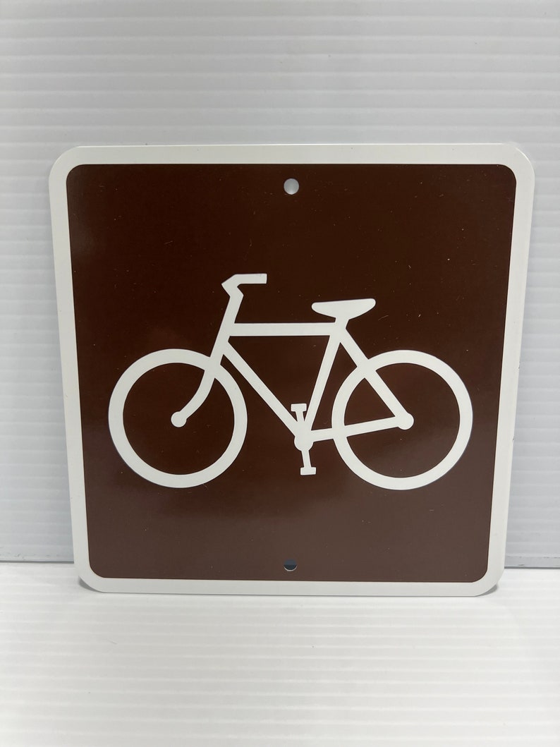 May include: A brown metal sign with a white bicycle symbol. The sign has two holes at the top and bottom.