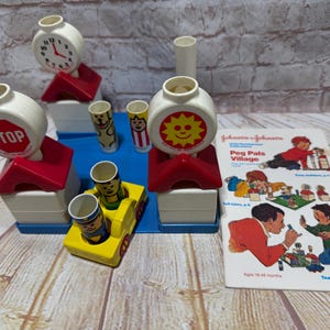 Könnte beinhalten: Ein farbenfrohes Spielzeugset aus Plastik für Kleinkinder, mit einer Uhr, einem Stoppschild und einer Sonne mit einem lächelnden Gesicht. Das Set enthält verschiedene Bausteine und Figuren. Die Schachtel enthält den Text "Peg Pals Village" und "Johnson & Johnson".