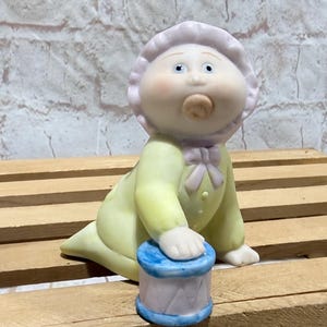 May include: A ceramic baby figurine in a yellow outfit and bonnet, with a pacifier, crawling towards a small drum. The drum is white with blue trim. The figurine has a light pink bow and bonnet.