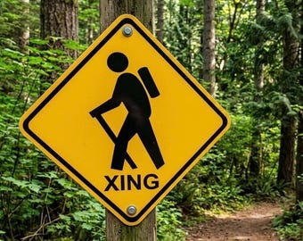 May include: A yellow diamond-shaped sign with a black hiker symbol and the word "XING" in black text. The sign is mounted on a wooden post in a forest setting with a hiking trail in the background.