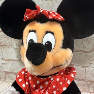 K&ouml;nnte beinhalten: Eine Minnie Mouse Pl&uuml;schpuppe mit gro&szlig;en schwarzen Ohren, einer roten Schleife mit wei&szlig;en Punkten und einem roten Kleid mit wei&szlig;en Punkten. Die Puppe hat eine schwarze Nase und gro&szlig;e Augen.