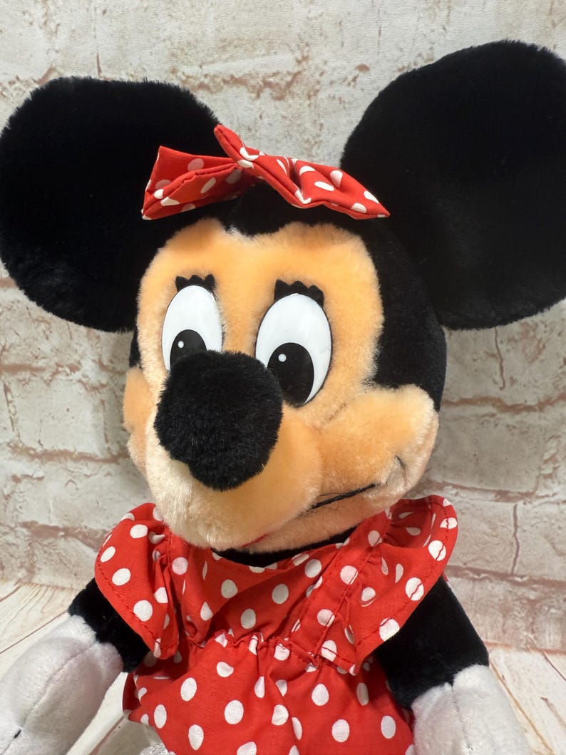 K&ouml;nnte beinhalten: Eine Minnie Mouse Pl&uuml;schpuppe mit gro&szlig;en schwarzen Ohren, einer roten Schleife mit wei&szlig;en Punkten und einem roten Kleid mit wei&szlig;en Punkten. Die Puppe hat eine schwarze Nase und gro&szlig;e Augen.