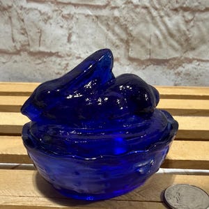 May include: A cobalt blue glass bunny figurine, featuring a detailed rabbit atop a bowl-shaped base. The figurine has a glossy finish and is set against a wooden surface. A coin is next to the figurine for size reference.