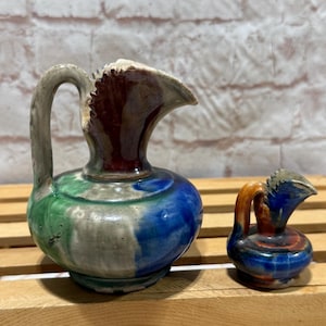 May include: Two small, decorative ceramic pitchers. The larger pitcher has a handle and is painted with blue, green, and gray hues. The smaller pitcher is a miniature version with similar colors. Both have a unique, textured rim.