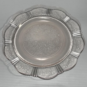 Puede incluir: Un plato de vidrio transparente y vintage con borde festoneado y un detallado diseño floral en el centro. El plato presenta detalles acanalados verticales alrededor del borde. El plato tiene aproximadamente 25 cm de diámetro.