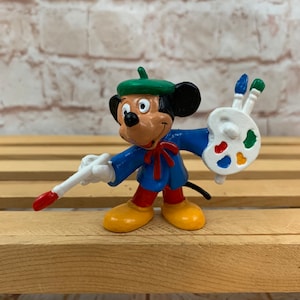 Może przedstawiać: Plastikowa figurka Myszki Miki przebranej za artystę. Ma na sobie niebieski fartuch, zielony beret i żółte buty. Trzyma pędzel i paletę z farbą.