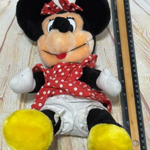 K&ouml;nnte beinhalten: Ein Pl&uuml;sch Minnie Mouse-Puppe mit schwarzen Ohren, einer rot-wei&szlig; gepunkteten Schleife und einem passenden Kleid. Die Puppe hat wei&szlig;e Handschuhe, schwarze Beine und gro&szlig;e gelbe F&uuml;&szlig;e. Ein Lineal liegt neben der Puppe.