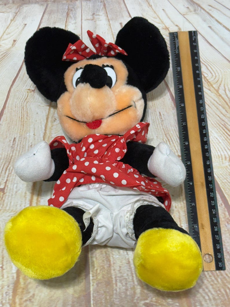 K&ouml;nnte beinhalten: Ein Pl&uuml;sch Minnie Mouse-Puppe mit schwarzen Ohren, einer rot-wei&szlig; gepunkteten Schleife und einem passenden Kleid. Die Puppe hat wei&szlig;e Handschuhe, schwarze Beine und gro&szlig;e gelbe F&uuml;&szlig;e. Ein Lineal liegt neben der Puppe.