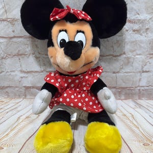 K&ouml;nnte beinhalten: Ein Minnie Mouse Pl&uuml;schtier. Die Puppe hat gro&szlig;e schwarze Ohren, eine rote Schleife mit wei&szlig;en Punkten und ein rotes Kleid mit wei&szlig;en Punkten. Die F&uuml;&szlig;e sind gelb und die H&auml;nde sind wei&szlig;. Ein klassischer Charakter.
