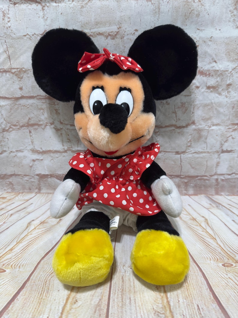K&ouml;nnte beinhalten: Ein Minnie Mouse Pl&uuml;schtier. Die Puppe hat gro&szlig;e schwarze Ohren, eine rote Schleife mit wei&szlig;en Punkten und ein rotes Kleid mit wei&szlig;en Punkten. Die F&uuml;&szlig;e sind gelb und die H&auml;nde sind wei&szlig;. Ein klassischer Charakter.