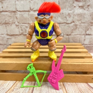 May include: A purple and yellow action figure of a troll with a red mohawk, wearing roller skates and holding a pink electric guitar. The figure is holding a green bow and arrow.