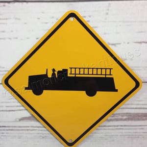 May include: Yellow diamond-shaped road sign with a black border. The sign depicts a black silhouette of a fire truck with a ladder.